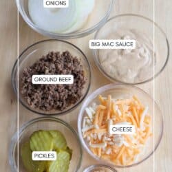 Six glass bowls on a wooden surface, each containing onions, Big Mac sauce, ground beef, shredded cheese, pickles, and sesame seeds.
