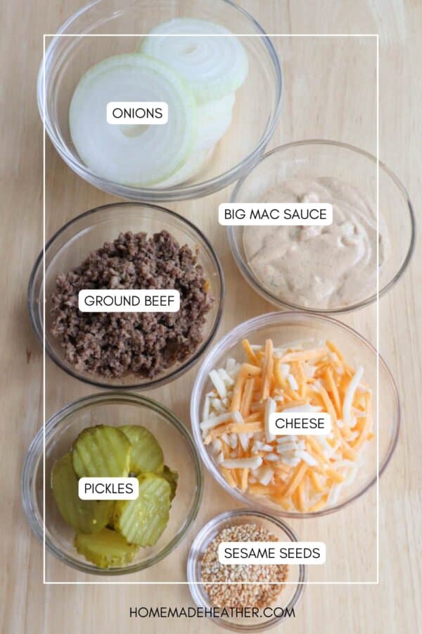 Six glass bowls on a wooden surface, each containing onions, Big Mac sauce, ground beef, shredded cheese, pickles, and sesame seeds.