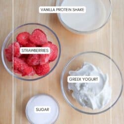 Top-down view of ingredients on a wooden surface: a bowl of vanilla protein shake, sliced strawberries, Greek yogurt, and a small bowl of sugar. Text labels identify each ingredient.
