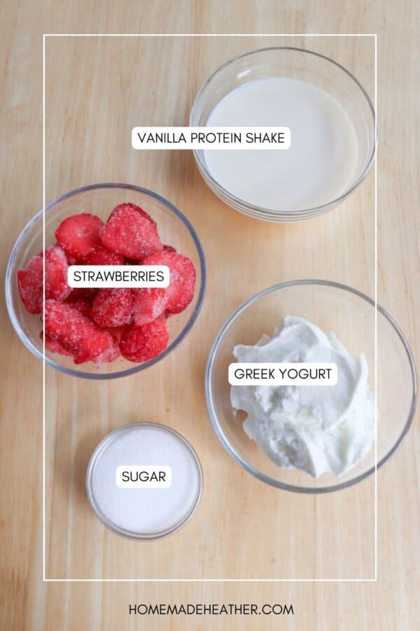 Top-down view of ingredients on a wooden surface: a bowl of vanilla protein shake, sliced strawberries, Greek yogurt, and a small bowl of sugar. Text labels identify each ingredient.