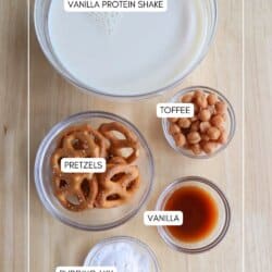 Top-down view of ingredients labeled for a vanilla protein shake: a bowl of liquid, a small bowl of toffee pieces, a bowl of pretzels, a small dish of vanilla extract, and a bowl of pudding mix on a wooden surface.