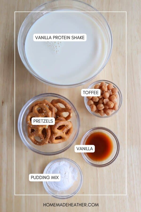 Top-down view of ingredients labeled for a vanilla protein shake: a bowl of liquid, a small bowl of toffee pieces, a bowl of pretzels, a small dish of vanilla extract, and a bowl of pudding mix on a wooden surface.