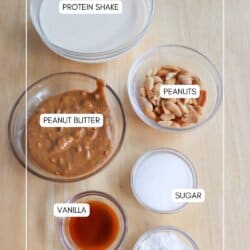 Overhead view of a wooden table with labeled ingredients: a bowl of protein shake, a bowl of peanuts, a bowl of peanut butter, a small bowl of vanilla, a small bowl of sugar, and a small bowl of pudding mix. The text homemadeheather.com is at the bottom.