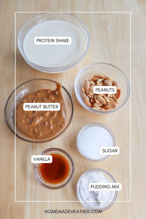 Overhead view of a wooden table with labeled ingredients: a bowl of protein shake, a bowl of peanuts, a bowl of peanut butter, a small bowl of vanilla, a small bowl of sugar, and a small bowl of pudding mix. The text homemadeheather.com is at the bottom.