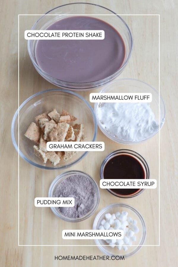 Top view of ingredients for a chocolate marshmallow dessert drink. Items include a bowl of chocolate protein shake, marshmallow fluff, graham crackers, chocolate syrup, pudding mix, and mini marshmallows, all labeled on a wooden surface.