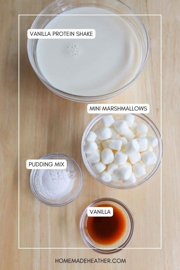 Four bowls with labeled ingredients on a wooden surface: Vanilla Protein Shake in a large bowl, Mini Marshmallows in a medium bowl, Pudding Mix in a small bowl, and Vanilla in a tiny bowl. Text reads homemadeheather.com.