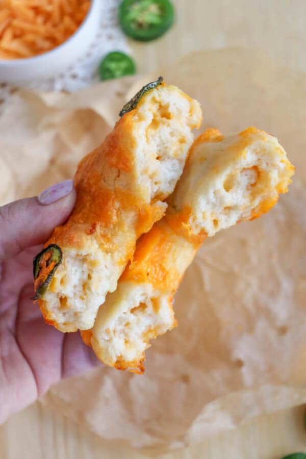 A hand holds two pieces of a cheese-stuffed, golden-brown biscuit or bread with jalapeรฑo slices on top. The bread appears soft and fluffy, with melted cheese visible. A bowl with shredded cheese and jalapeรฑo slices is in the background.