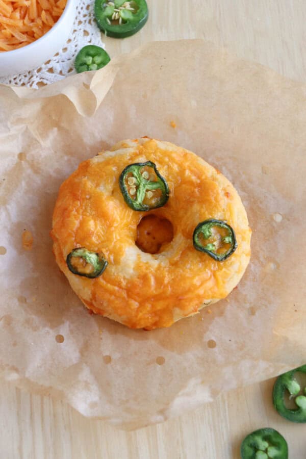 A bagel topped with melted cheese and sliced jalapeรฑos sits on a piece of parchment paper. In the background, there is a bowl with shredded cheese and scattered jalapeรฑo slices on a wooden surface.