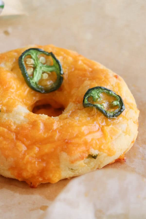 A bagel topped with melted cheddar cheese and sliced jalapeรฑos, resting on parchment paper.