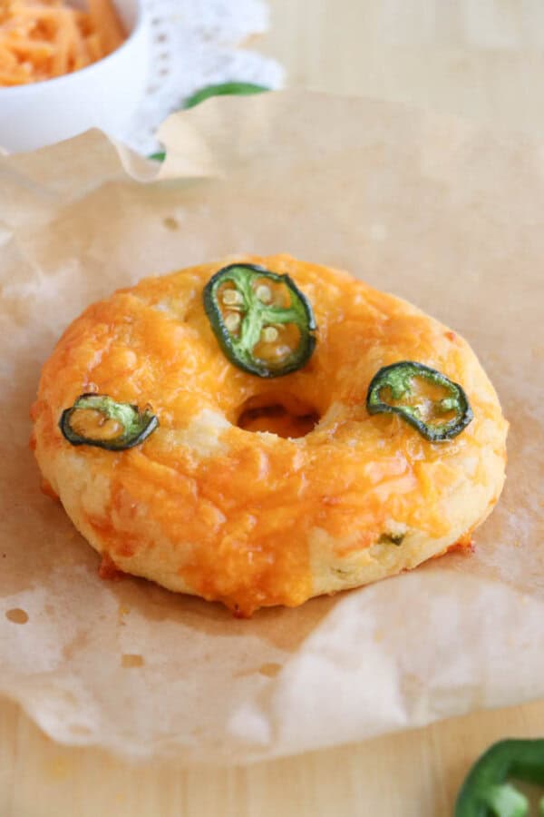 A bagel topped with melted cheddar cheese and sliced jalapeรฑos sits on brown parchment paper. The bagel appears savory and spicy, with green jalapeรฑo slices adding a pop of color against the golden cheese.