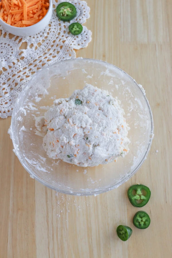 A glass bowl with dough containing green and orange bits sits on a wooden table. Nearby are sliced jalapeรฑos and shredded orange cheese in a small round dish on a lace cloth.