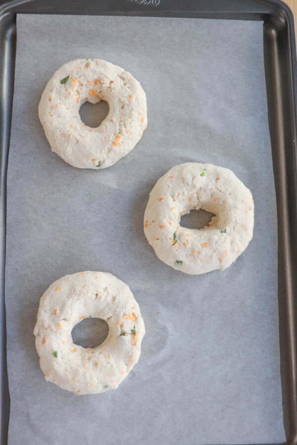 Three unbaked, circular bagels with bits of vegetables are placed on a parchment-lined baking sheet.