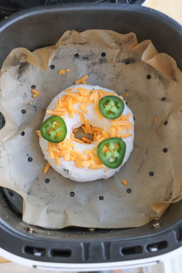 A bagel topped with shredded cheese and three jalapeรฑo slices is placed on a piece of parchment paper inside an air fryer basket. The bagel appears uncooked, with a white doughy texture.