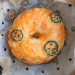 A bagel topped with melted cheese and three slices of jalapeño, sitting on parchment paper with small holes. The bagel is golden brown and appears freshly baked.