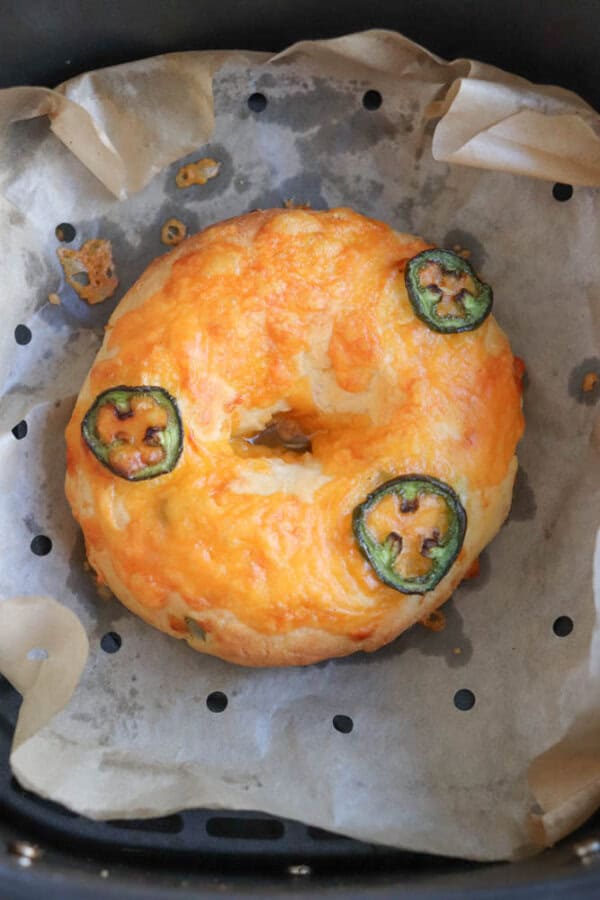 A bagel topped with melted cheese and three slices of jalapeรฑo, sitting on parchment paper with small holes. The bagel is golden brown and appears freshly baked.