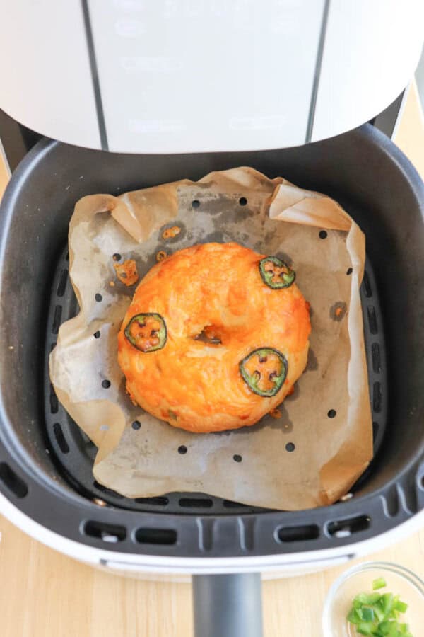A bagel topped with melted cheese and jalapeรฑo slices sits on parchment paper inside an air fryer basket. The parchment is slightly burnt at the edges. A small dish of green garnish is partially visible in the corner.