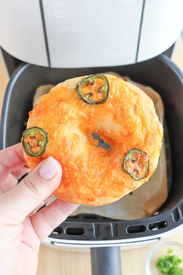 A hand holds a freshly cooked bagel topped with melted cheese and slices of jalapeรฑo. The bagel is being lifted from the basket of an air fryer. A small dish with green herbs is partially visible in the background.