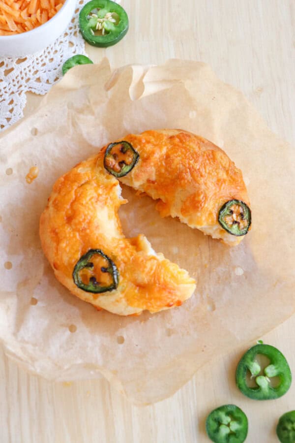 A cheesy pastry with a bite taken out, topped with jalapeรฑo slices, rests on parchment paper. Shredded cheese and jalapeรฑo slices are scattered around it on a wooden surface.