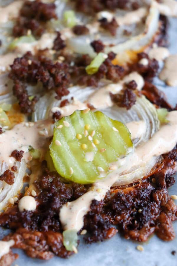 Close-up of a cheeseburger topped with crumbled ground beef, melted cheese, sliced onions, a pickle, and drizzled sauce. The toppings are spread over a toasted brown patty, emphasizing textures and layers.