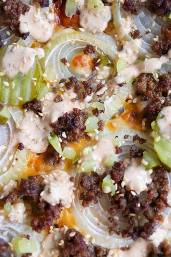Close-up of a pizza topped with cheese, seasoned beef crumbles, sliced pickles, diced onions, sesame seeds, and drizzled with a creamy sauce. The toppings are spread across a thin crust.