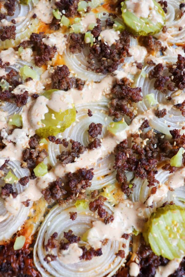 Close-up of a casserole topped with sliced onions, crumbled ground meat, pickles, and a drizzle of creamy sauce. The surface is browned and slightly crispy.