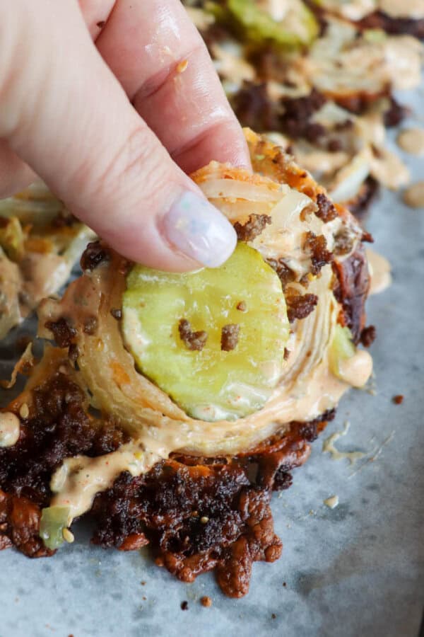Close-up of a hand holding a crispy fried pickle chip topped with melted cheese and a slice of pickle, drizzled with sauce, on a parchment-lined surface. The chip has a golden-brown crust and is part of a group of similar snacks.