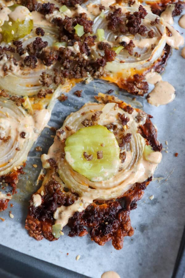 Close-up of a cheeseburger pizza with toppings including crumbled ground beef, pickles, onions, and a drizzle of special sauce, set on a baking sheet lined with parchment paper. A slice is slightly pulled away from the rest.