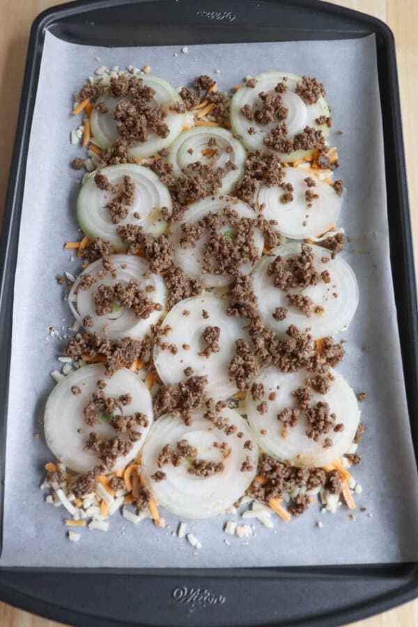 Slices of onion topped with ground meat and shredded cheese are arranged on a parchment-lined baking tray.