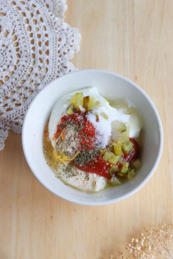 A white bowl on a wooden table contains mayonnaise, ketchup, diced pickles, and various spices. A doily is partially visible to the side.