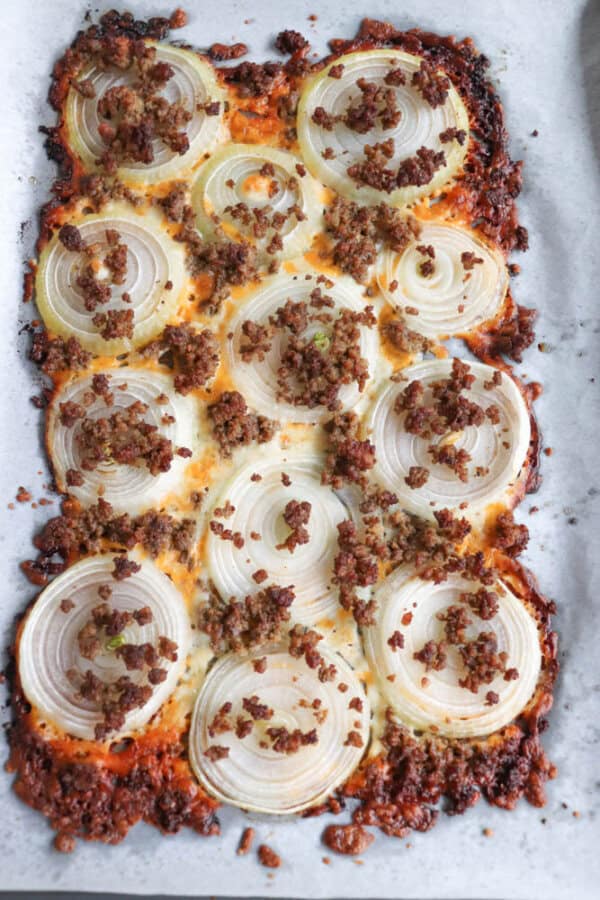 A baked dish on parchment paper featuring a layer of sliced onions topped with ground meat, likely beef or sausage. The edges are crispy and browned, suggesting it is freshly baked. The round onion slices are evenly distributed across the dish.