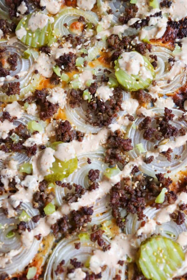 Close-up of a pizza topped with sliced onions, pickles, ground beef, and drizzled with a creamy sauce. The toppings create a textured look with a variety of colors and flavors on a golden-brown crust.