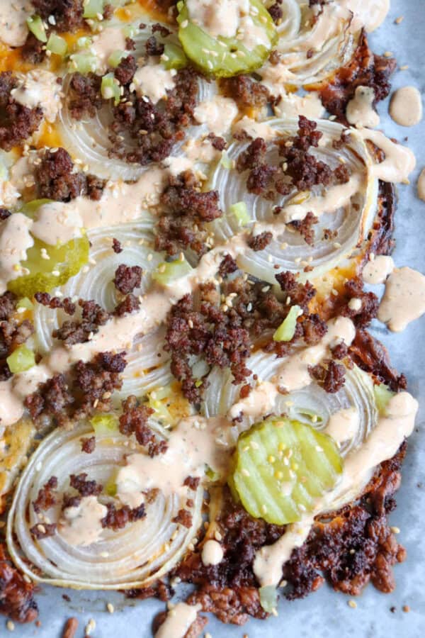 Close-up of a loaded pizza topped with sliced onions, crumbled sausage, pickle slices, and drizzled with a creamy sauce. The crust appears crispy and golden brown, with a generous layer of toppings.