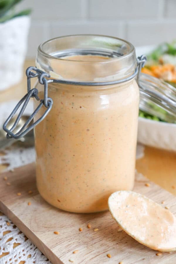 A glass jar of creamy orange sauce with herbs inside, placed on a wooden board. A spoon with sauce rests beside the jar. A blurred salad is in the background.