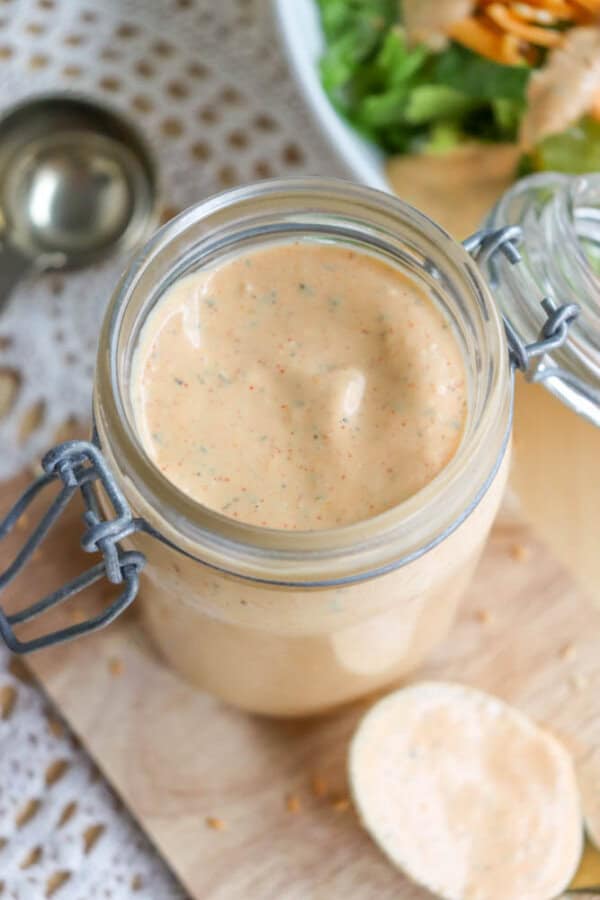 A jar of creamy, orange-colored dressing with visible specks, placed on a wooden board. A spoon rests nearby. A partial view of a salad bowl is in the background.