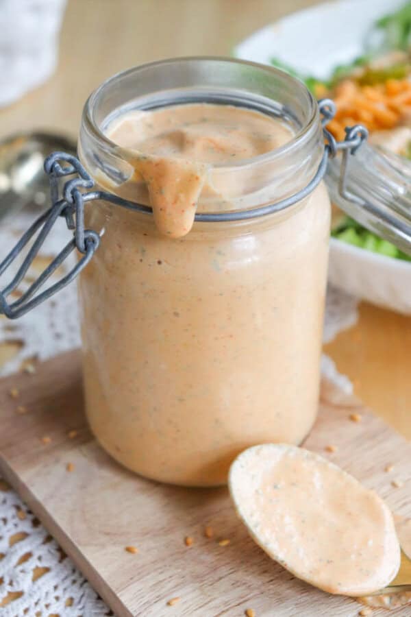 A glass jar filled with creamy orange dressing, slightly overflowing and dripping down the side. The jar sits on a wooden board, accompanied by a spoon with dressing on it. A salad is blurred in the background.