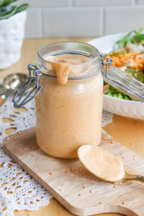 A jar of creamy orange sauce sits on a wooden board, with a spoonful of the sauce resting beside it. The sauce drips slightly from the jar. In the background, theres a plate of fresh salad and a blurred white brick wall.