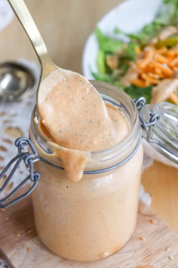 A spoon is lifting creamy dressing from a glass jar with a hinged lid. The dressing is light orange with specks of herbs. In the background, there is a partially visible green salad with shredded carrots on a white plate.