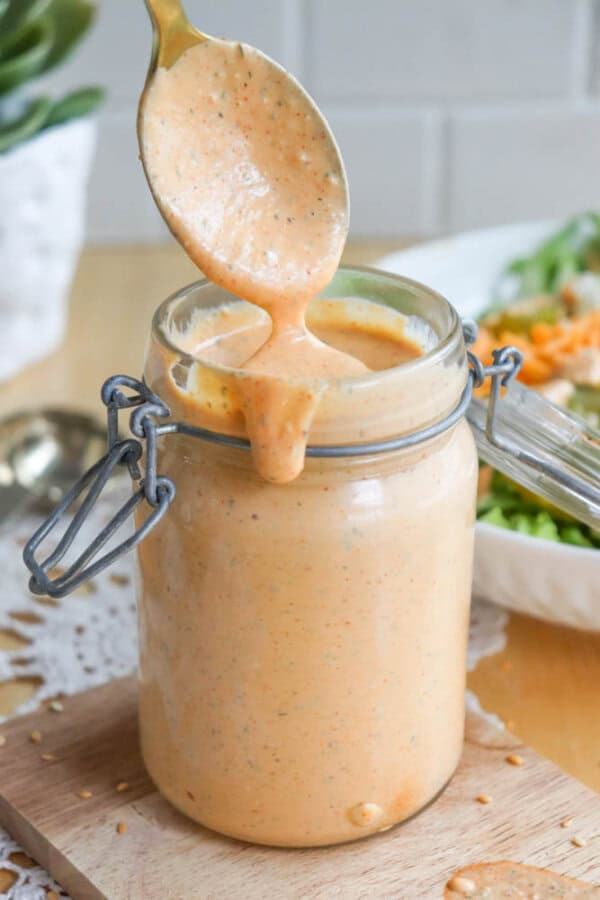 A spoon drizzles a creamy orange sauce into an open glass jar on a wooden board. The sauce has black specks and is spilling slightly over the jars edge. A salad and a spoon are blurred in the background.