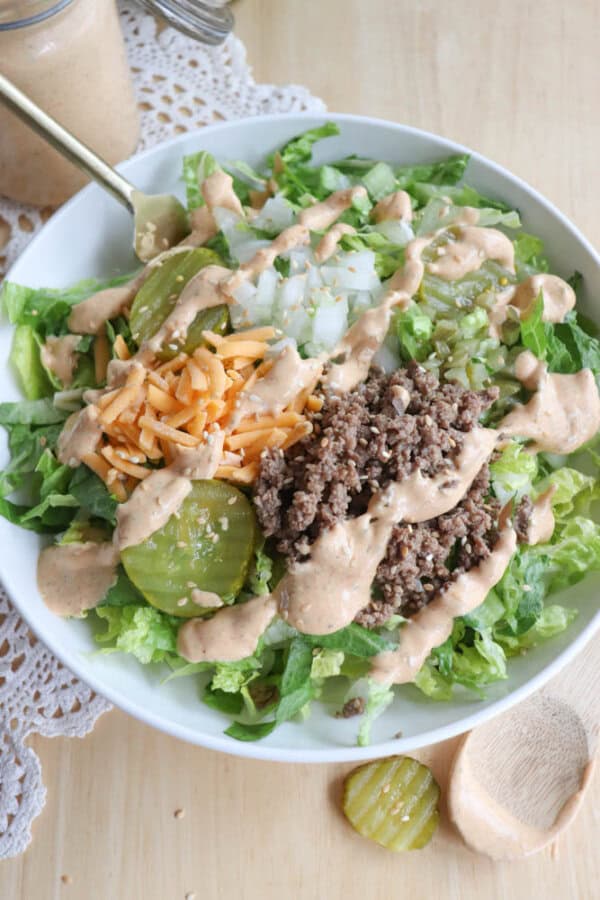 A deconstructed burger salad in a white bowl featuring chopped lettuce, ground beef, shredded cheddar cheese, diced onions, pickles, and a creamy dressing drizzled on top. A wooden spoon and a jar of dressing are beside the bowl.