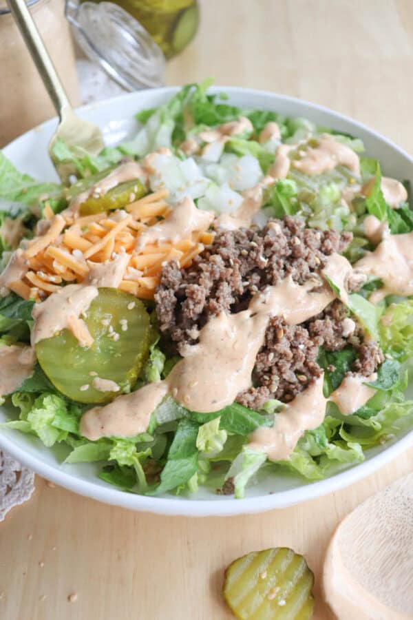A salad topped with cooked ground beef, shredded cheddar cheese, diced onions, pickles, and a creamy dressing on fresh lettuce. A fork and a jar of pickles are visible in the background.