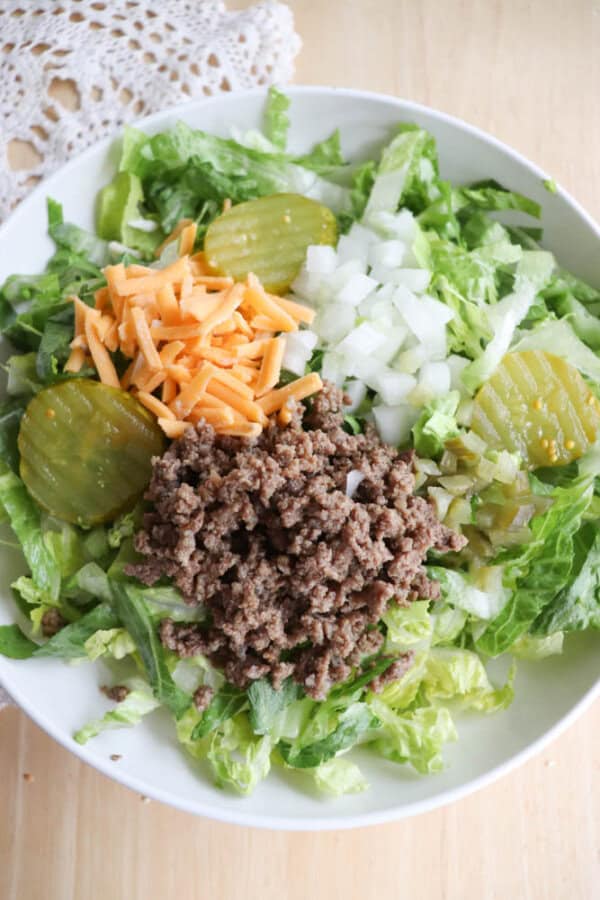 A salad in a white bowl featuring shredded lettuce, cooked ground beef, sliced pickles, diced onions, and shredded cheddar cheese. A lace doily is partially visible in the background.