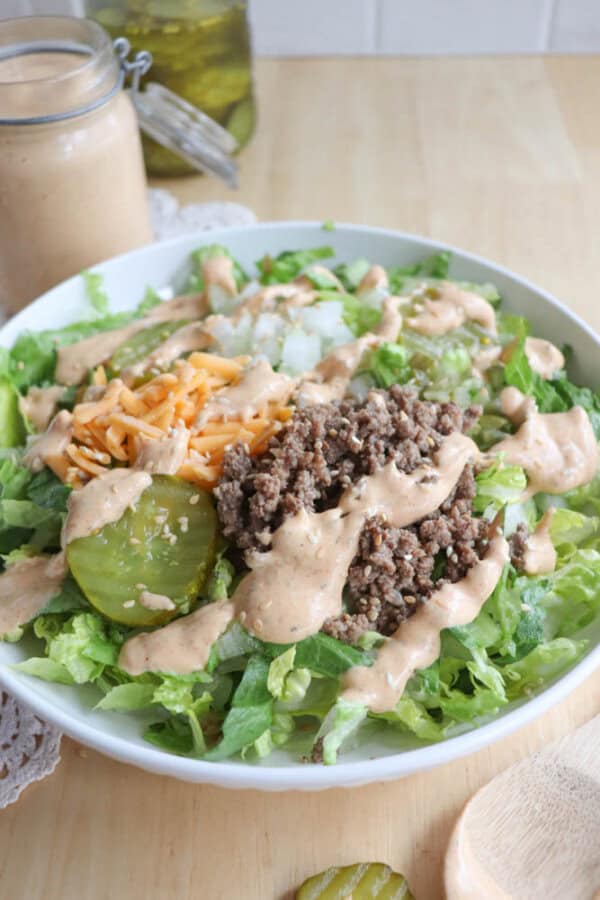 A salad in a white bowl featuring beef, shredded cheddar cheese, pickles, diced onions, and chopped lettuce, topped with a creamy dressing. A jar of dressing and a glass jar of pickles are in the background.
