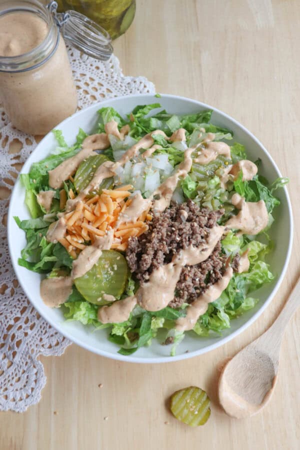 A salad in a white bowl on a wooden table, topped with ground meat, shredded cheese, chopped onions, sliced pickles, and drizzled with creamy dressing. A jar of dressing and a wooden spoon are nearby, with a lace doily adding a decorative touch.
