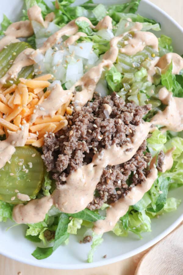 A bowl of salad containing chopped lettuce, shredded cheddar cheese, diced onions, pickles, crumbled ground beef, and a drizzle of creamy dressing.