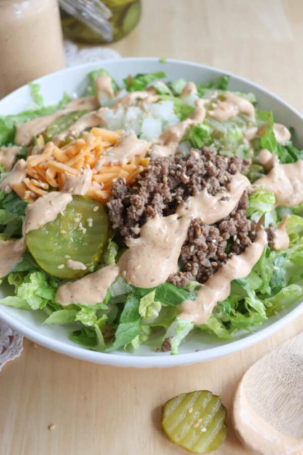 A salad topped with ground beef, shredded cheese, pickles, and a creamy dressing sits on a white plate. The bed consists of fresh lettuce. A jar of dressing and a pickle slice are in the background on a wooden surface.