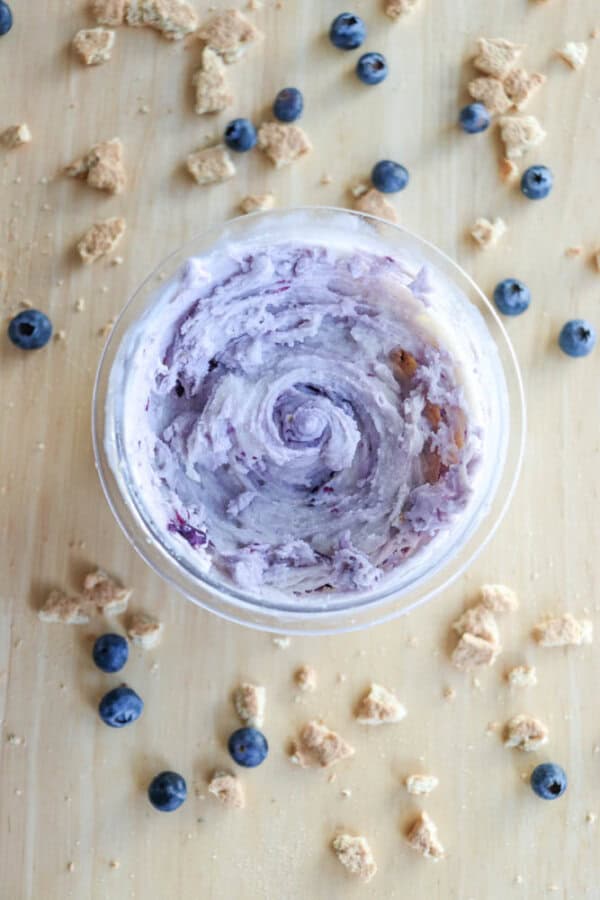 A bowl filled with swirled blueberry cheesecake ice cream is placed on a wooden surface. Surrounded by scattered blueberries and crumbled cookies, the creamy dipโreminiscent of blueberry cheesecake ice creamโappears light purple with rich texture.