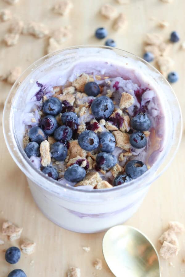 A cup filled with blueberry ice cream, reminiscent of blueberry cheesecake ice cream, is topped with fresh blueberries and crushed granola. A gold spoon rests nearby on a wooden surface, surrounded by scattered blueberries and granola pieces.