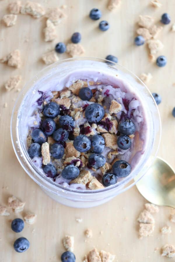 A bowl of ice cream topped with fresh blueberries and crumbled granola sits on a wooden surface, reminiscent of blueberry cheesecake ice cream. Additional blueberries and granola bits are scattered around, while a gold spoon is partially visible to the right of the bowl.