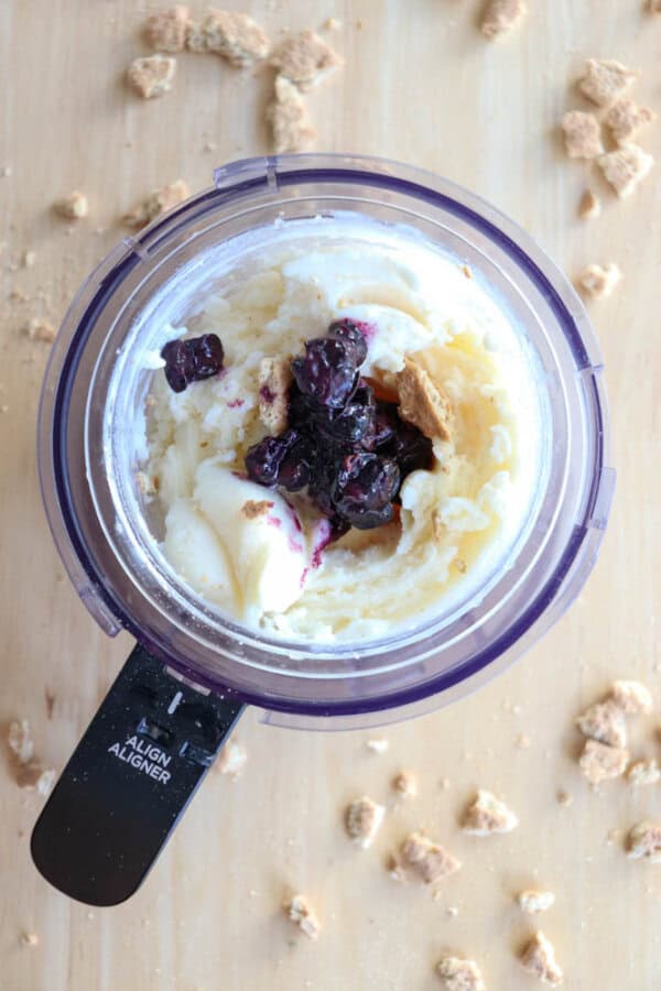 A food processor containing creamy blueberry cheesecake ice cream swirled with dark berry sauce and crushed cookie pieces. Scattered cookie crumbs are visible on the light wooden surface around the processor.