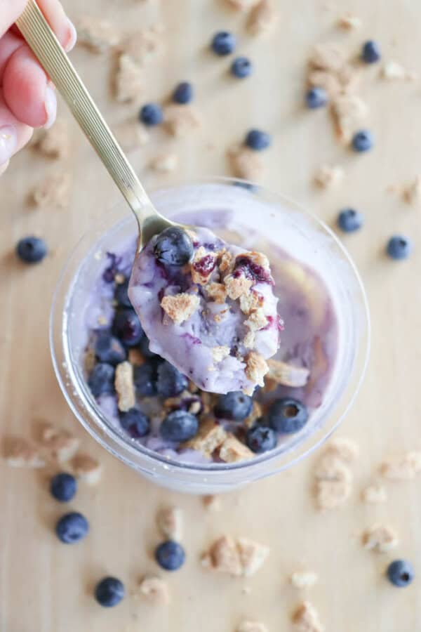 A hand holds a spoonful of ice cream reminiscent of blueberry cheesecake ice cream, topped with fresh blueberries and crunchy granola, over a jar brimming with the same creamy mixture. Blueberries and granola pieces are artfully scattered on the light wooden surface around the jar.